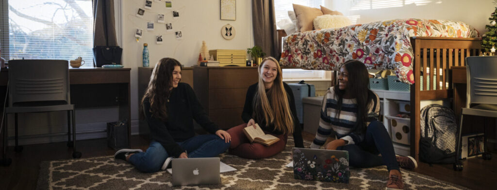 Three students sitting in a dorm room looking at laptops
