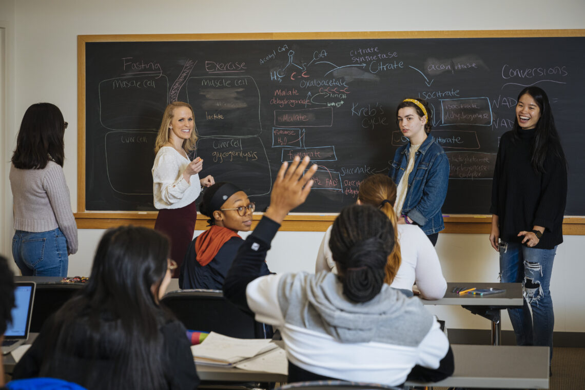 Students and Professor at a chalkboard while other students raise hands to answer a question.