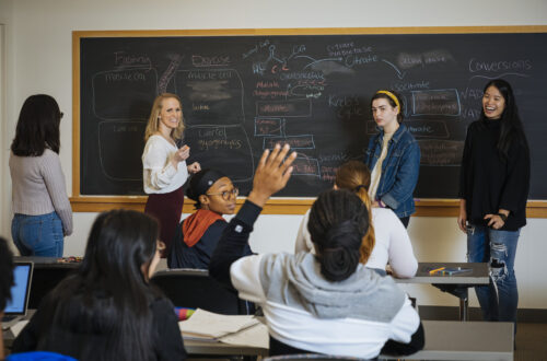 Students and Professor at a chalkboard while other students raise hands to answer a question.