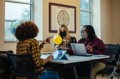 Two students gather around laptops and a model of the solar system in discussion.