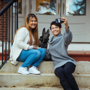Two students sit on the steps of Rebekah Scott Hall. They are taking a selfie with Ramona, a statue of a Scottie dog.