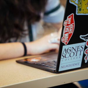 Unseen student sits behind a laptop with numerous stickers, including one that reads "Agnes Scott College Neuroscience"