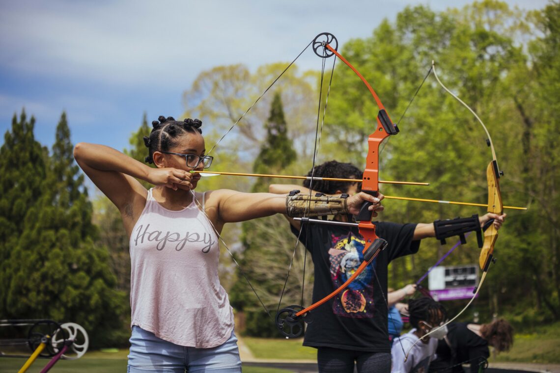 Students in a line aim at a target with a bow and arrowl