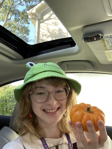Leah - pictured smiling in her glasses with a bright green, frog hat - holding a pumpkin with pinked polished nails sitting in the car.