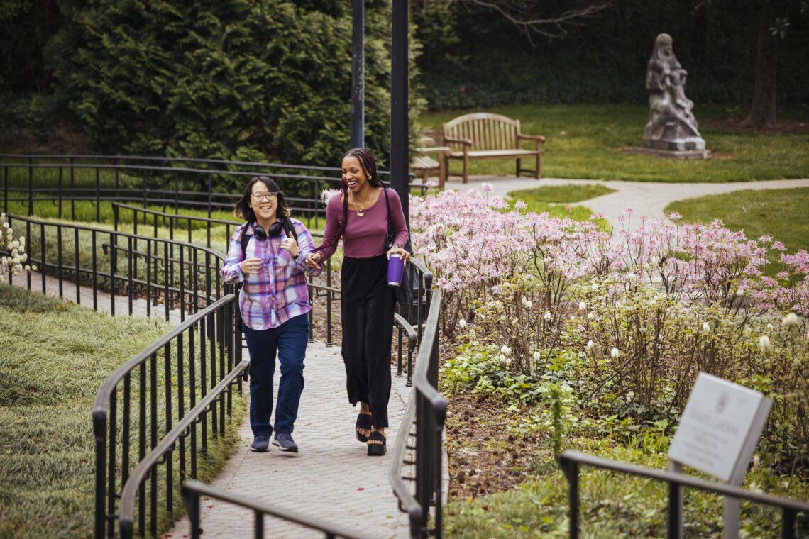 Two students walk and talk up a pathway surrounded by spring flowers.