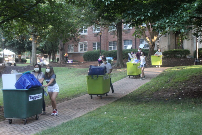 Several students and families wearing masks push large green bins full of dorm room items.