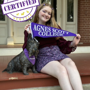 Student with a maroon shirt, purple skirt, and cat-eye eyeliner holds a purple pennant reading "Agnes Scott College" while sitting next to Scottie dog statue.