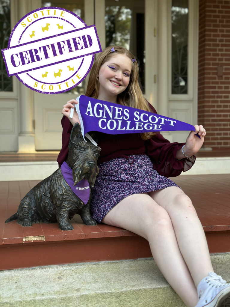 Student with a maroon shirt, purple skirt, and cat-eye eyeliner holds a purple pennant reading "Agnes Scott College" while sitting next to Scottie dog statue.