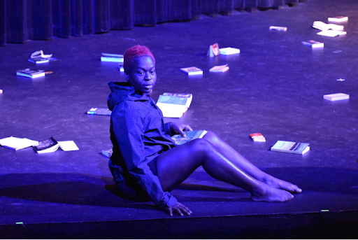 Dancer sits on stage, surrounded by books strewn across the ground.