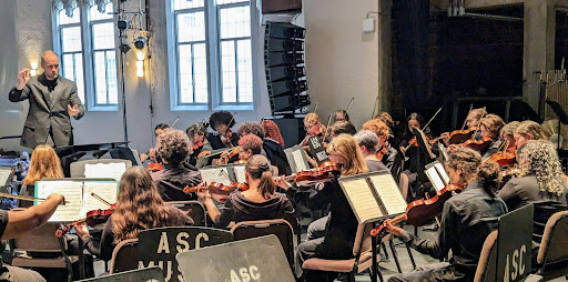 View of conductor and behind performers of string orchestra.
