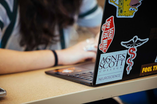 Unseen student sits behind a laptop with numerous stickers, including one that reads "Agnes Scott College Neuroscience"
