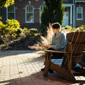 A student sits in a wooden adirondack chair while looking down at a folder.