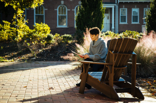 A student sits in a wooden adirondack chair while looking down at a folder.